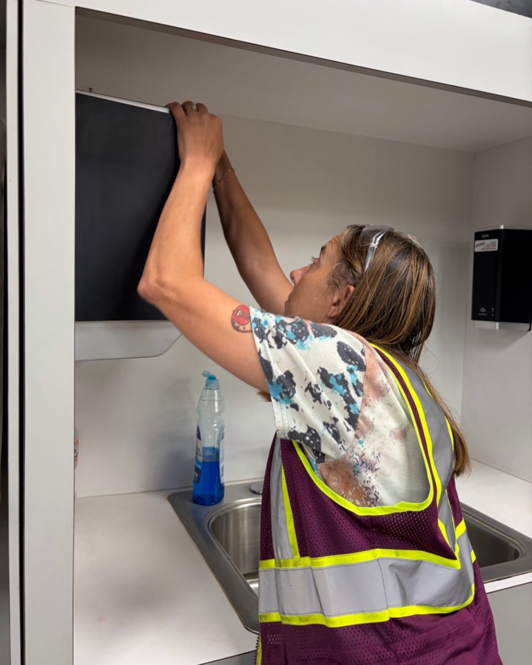 An HM Solutions crew member cleaning inside a canteen cabinet above a breakroom sink