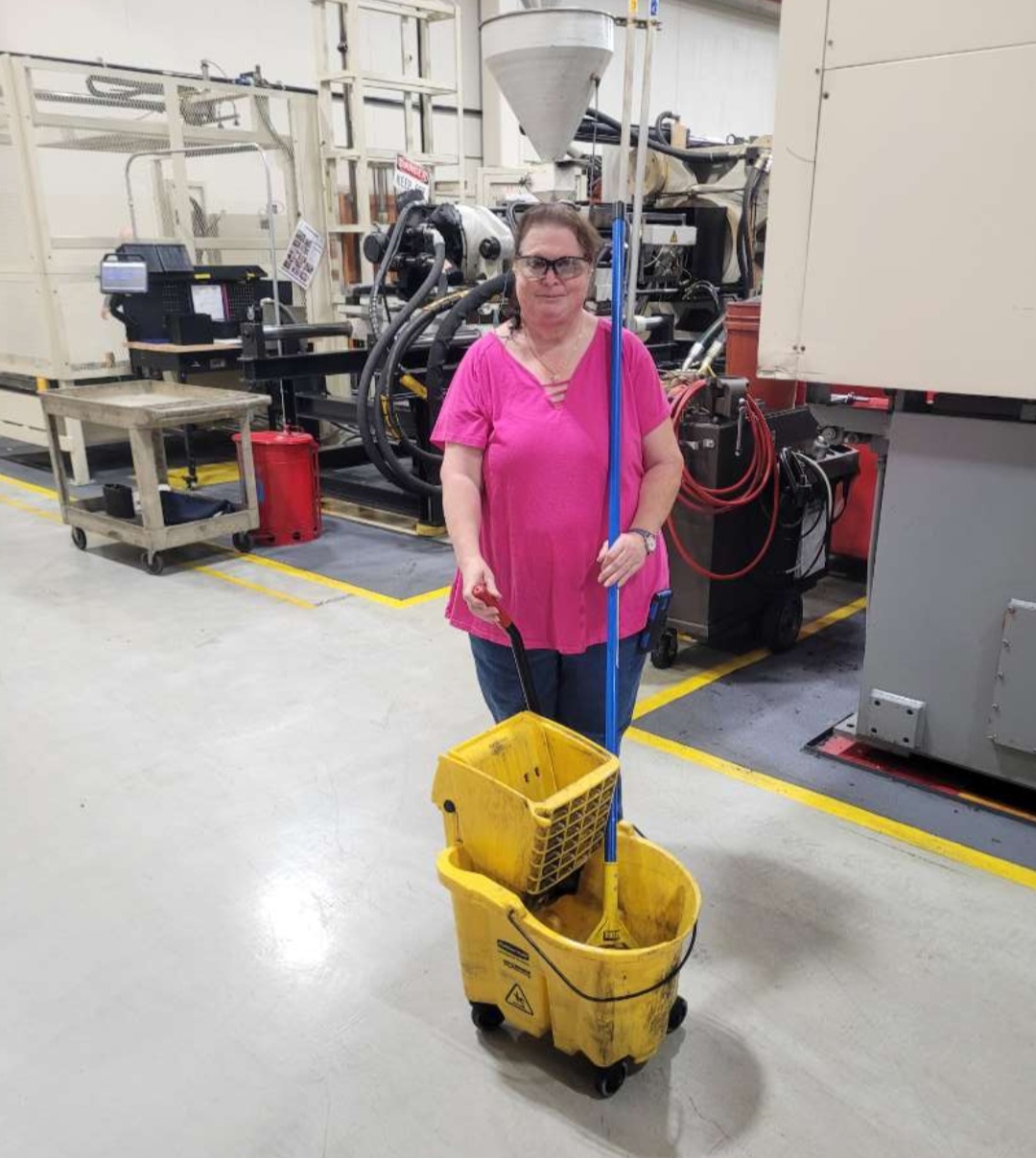 An HM Solutions crew member with a mop and bucket on a production floor beside injection molding equipment